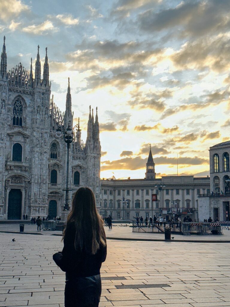 Woman with long hair seen from behind standing in a large square, facing the Milan Cathedral's ornate Gothic facade at sunset.