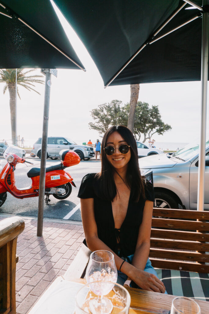 Smiling woman in a black plunging top and round sunglasses sits at an outdoor seaside cafe table under a large umbrella, with a red scooter and parked cars behind her.
