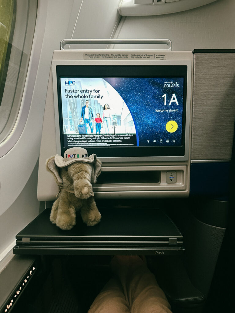 In-flight entertainment screen mounted on the seatback with a plush gray elephant toy resting on the tray table in front of it.