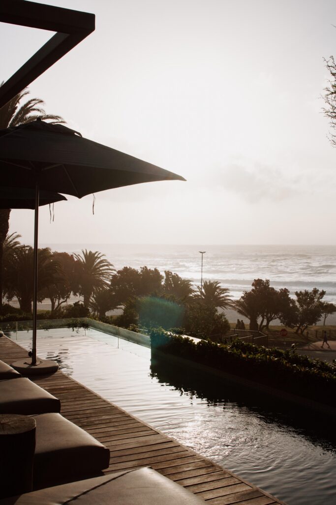 Resort pool with a wooden deck and a large umbrella overlooking palm trees and a sunlit ocean.