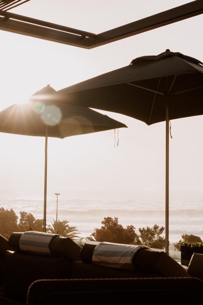 Sunlit outdoor lounge with large patio umbrellas overlooking the ocean and distant palm trees.