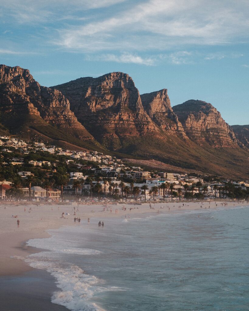 White sandy beach with light waves, a row of palm trees, and a mountain town at the base of rugged cliffs under a blue sky.