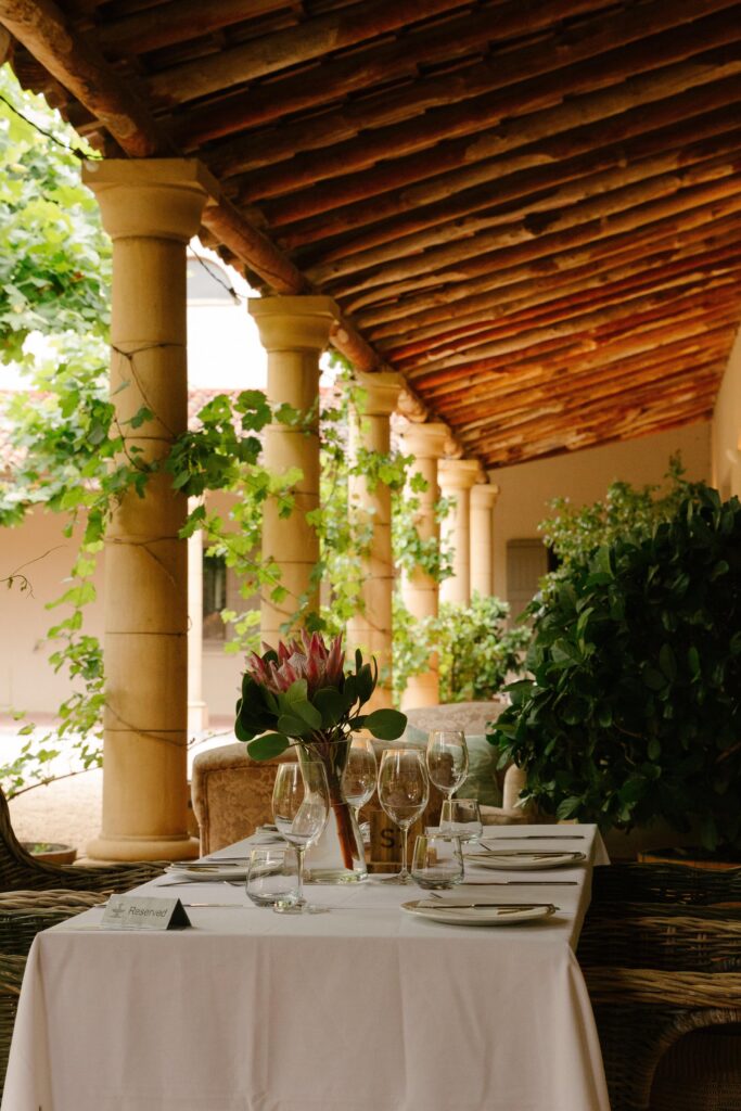 Outdoor dining table set on a vine-covered terrace with glassware and a botanical centerpiece under a tiled pergola.