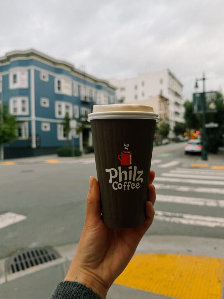 Hand holding a Philz Coffee cup in foreground with a blue building and street scene blurred in the background.