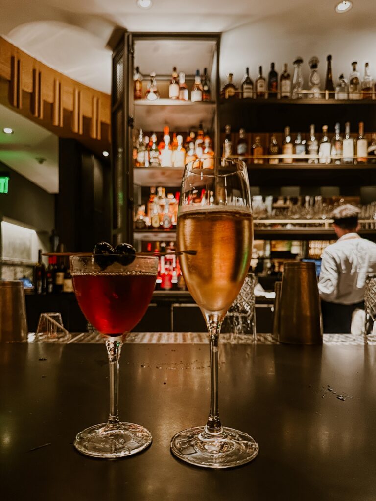 Two stemmed wine glasses on a dark bar counter with a backdrop of bottles on shelves in a warm, dimly lit bar.