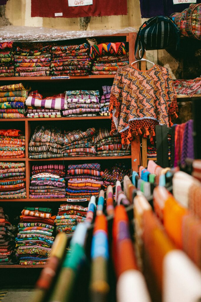 Vibrant market stall with shelves of folded, patterned textiles and an orange fringed shawl hanging in front.