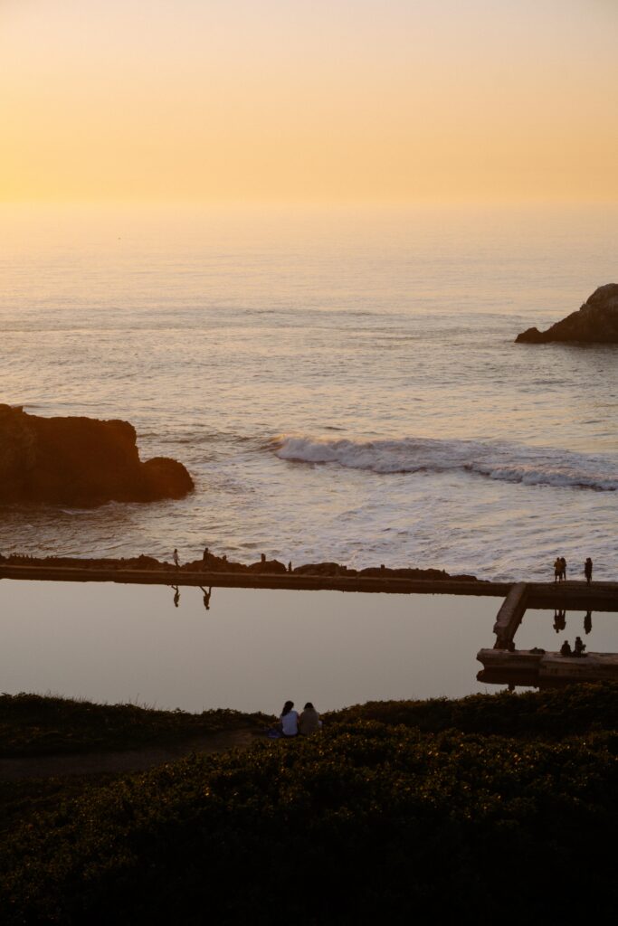 Golden sunset over a calm sea with rocky shores in the foreground; a couple sits on the grassy edge by the water's reflection.
