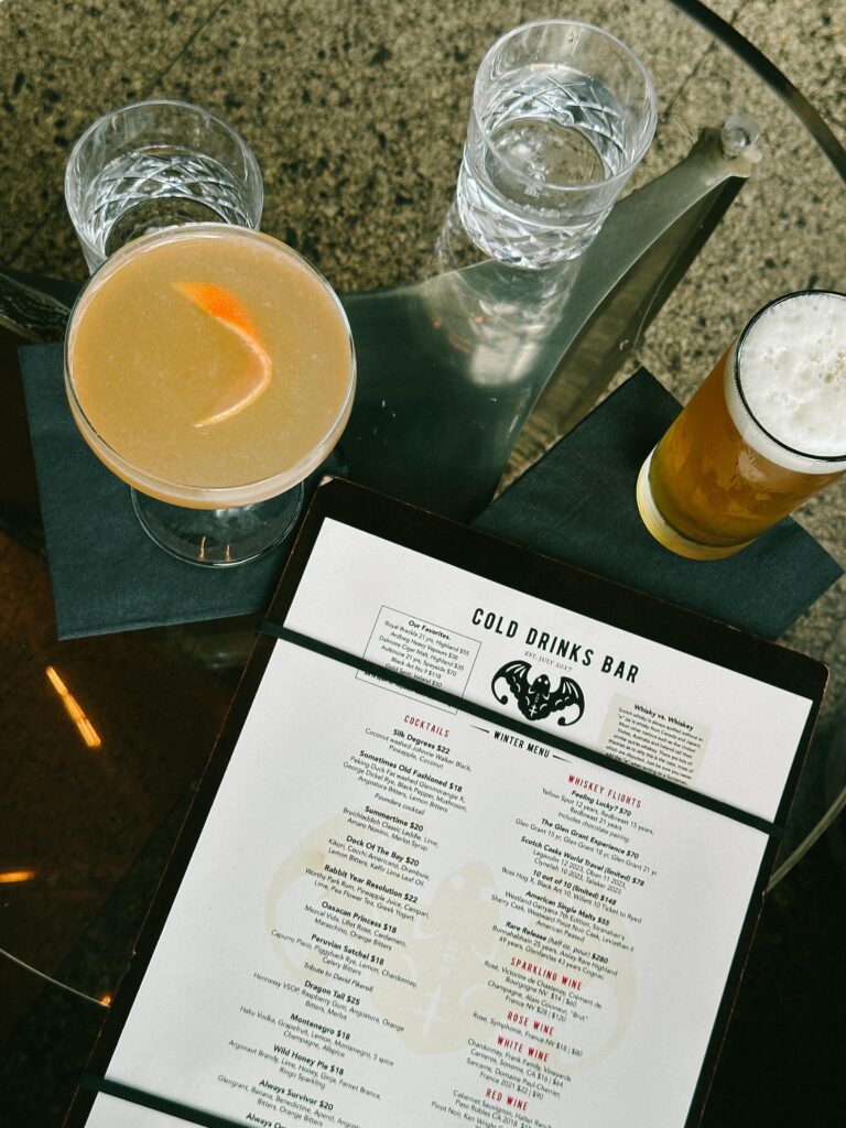 Pale cocktail with orange twist on a table, flanked by two crystal water glasses and a frothy beer nearby beside a menu.