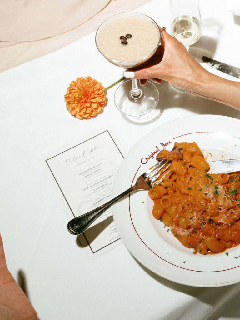 A hand lifts a cocktail glass over a plate of orange pasta on a white table, with a bright orange flower and a menu nearby.