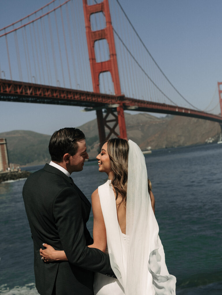 A bride and groom embrace by the water with the Golden Gate Bridge in the background, on their wedding day.