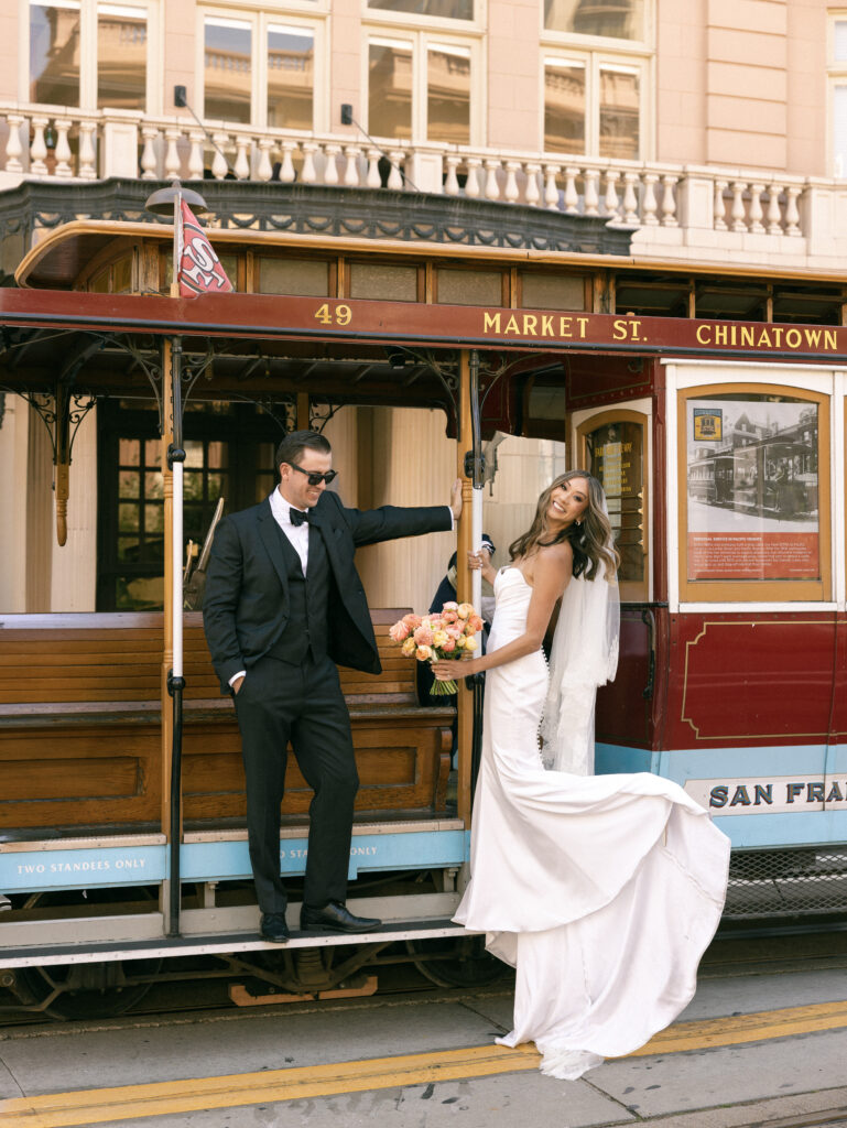 Bride in a white gown and groom in a black tux pose on a vintage Market Street trolley, Chinatown; she holds a peach-pink bouquet.