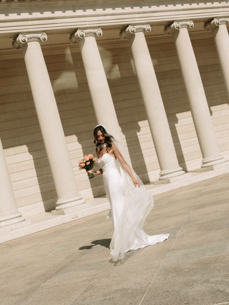 Bride in a white wedding gown holding a pink bouquet walks between tall classical columns of a neoclassical building.