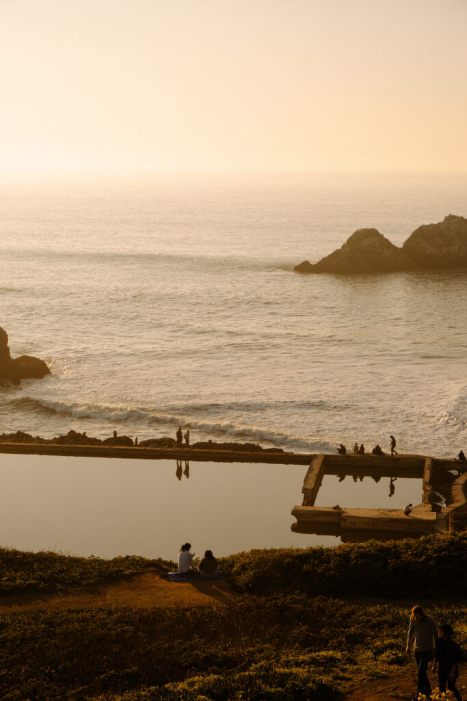 Golden sunset over a calm sea with rocky outcrops; people sit and stand along the shore, and a reflective pool sits in the foreground on the right.