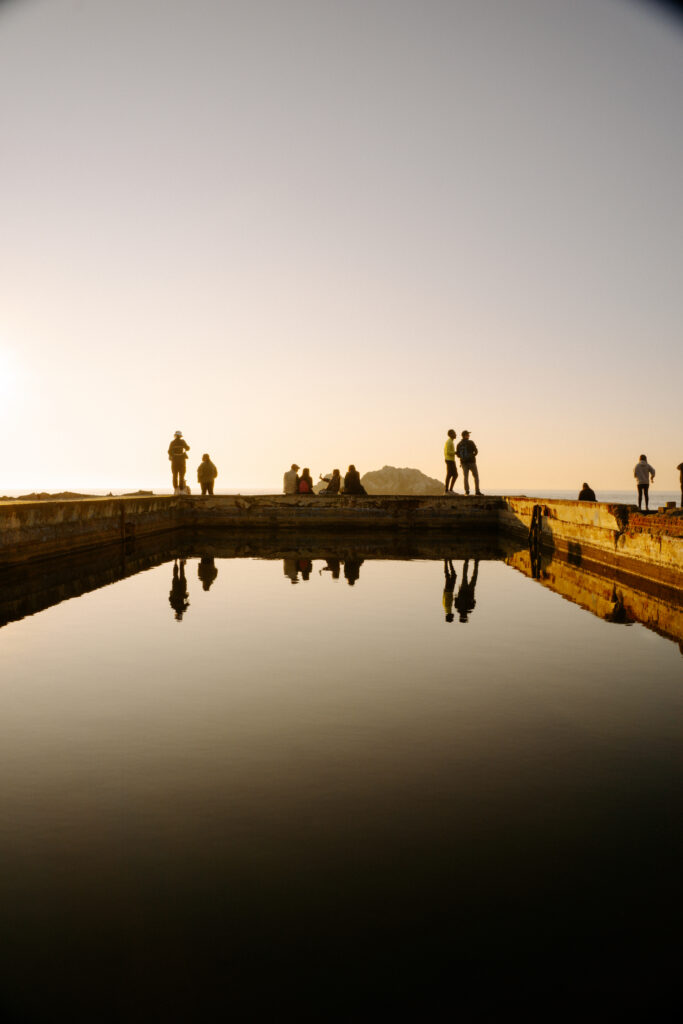 Group of people along a wooden pier at sunset, with calm water and their reflections visible in the pool below.