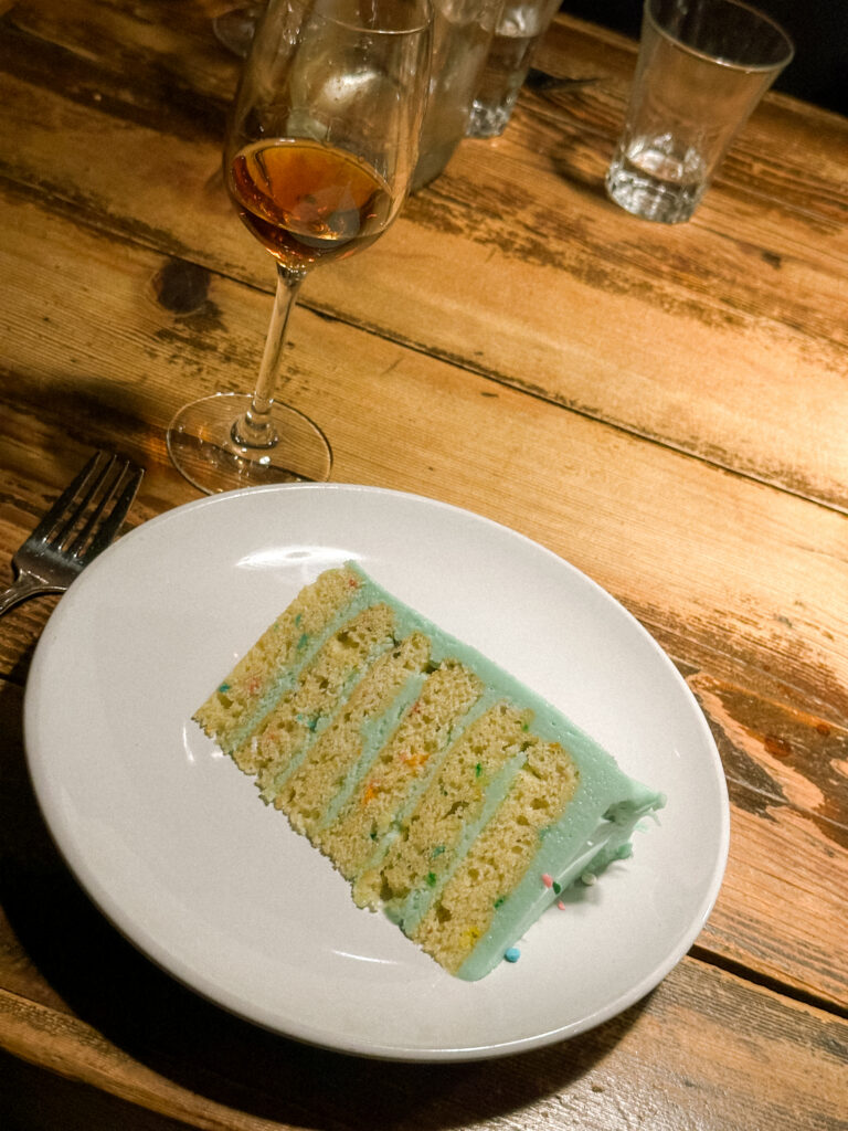 Slice of layered cake with light blue frosting on a white plate, next to a fork on a rustic wooden table, with a wine glass in the background