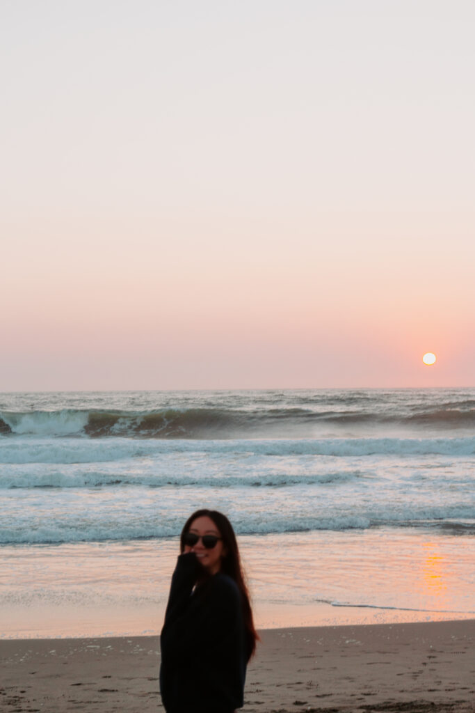 Person wearing sunglasses stands on a sandy beach at sunset, with gentle waves and a pale pastel sky behind them.