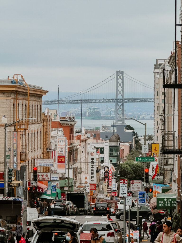 Busy Chinatown street filled with storefronts, colorful signs, pedestrians, and cars, with a distant suspension bridge in the background.