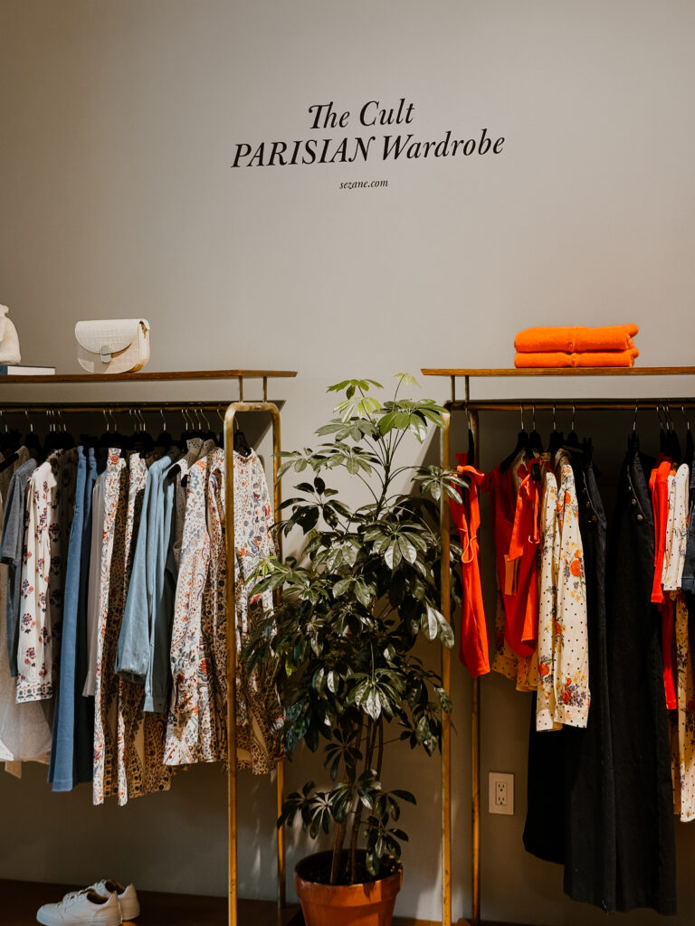 Display of a boutique with hanging floral and denim dresses on left and bright orange outfits on the right, a potted plant between racks, and a white handbag on the left shelf.