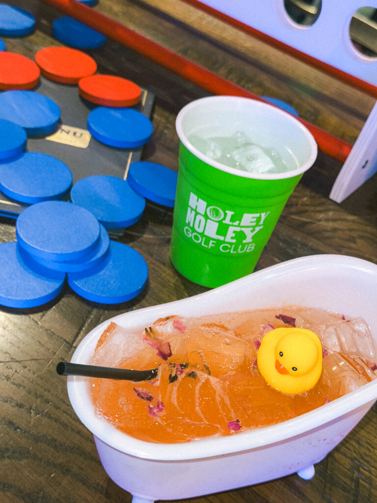 Green cup with ice water labeled Holy Moley Golf Club, beside a tub of orange punch and a yellow rubber duck, with blue and orange game chips nearby on a wooden table.