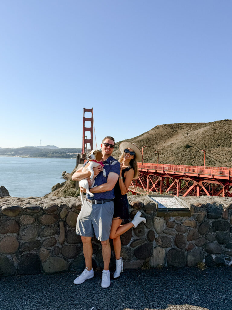Couple holding a small dog by a stone wall with the Golden Gate Bridge in the background on a bright day.
