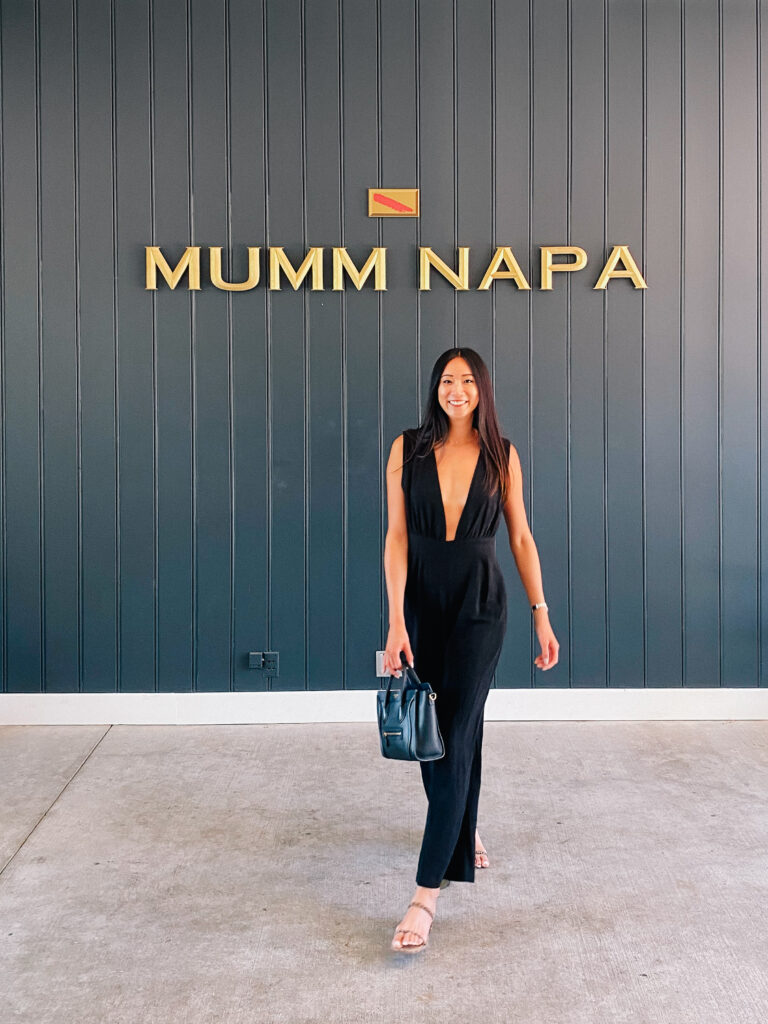 Smiling woman in a black sleeveless jumpsuit with a blue handbag stands in front of a dark paneled wall with a gold MUMM NAPA sign above her.