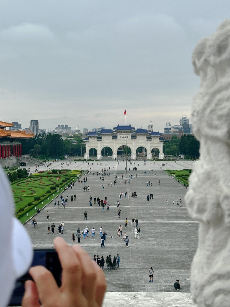 Wide, open plaza with a white arch gateway topped with blue roofs and a tall flagpole in the center; distant city skyline visible; hands and a phone in the foreground indicate a tourist photo.