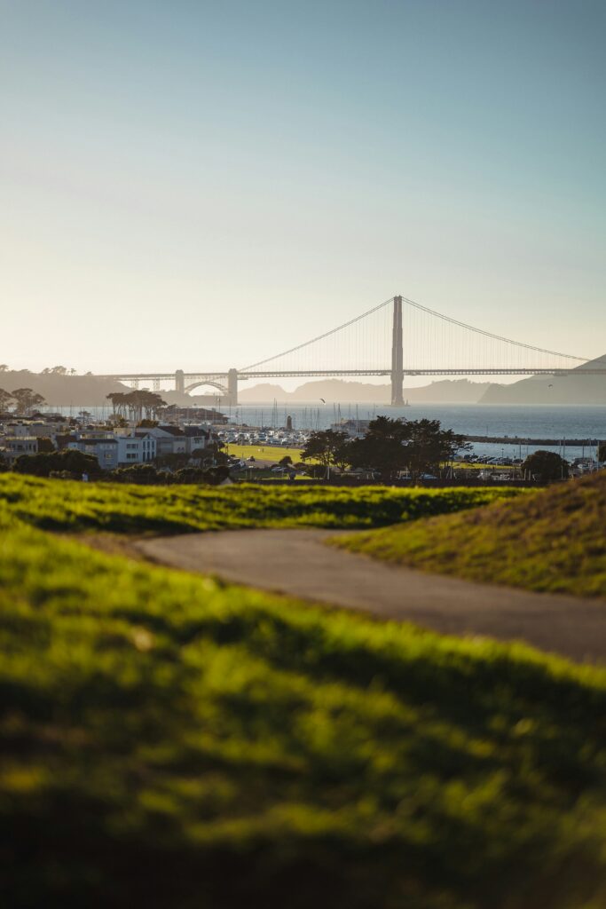 Suspension bridge spanning a calm bay with a coastal town and green grassy foreground beneath a pale blue sky at sunset/sunrise.