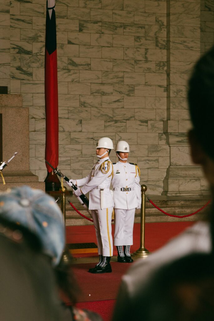 Two ceremonial guards in white uniforms and helmets stand at attention on a red carpet beside a flagpole inside a stone-walled hall.