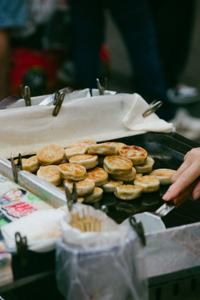 Pile of small round fried pastries cooking on a griddle at a street stall, with metal clips and paper lining nearby