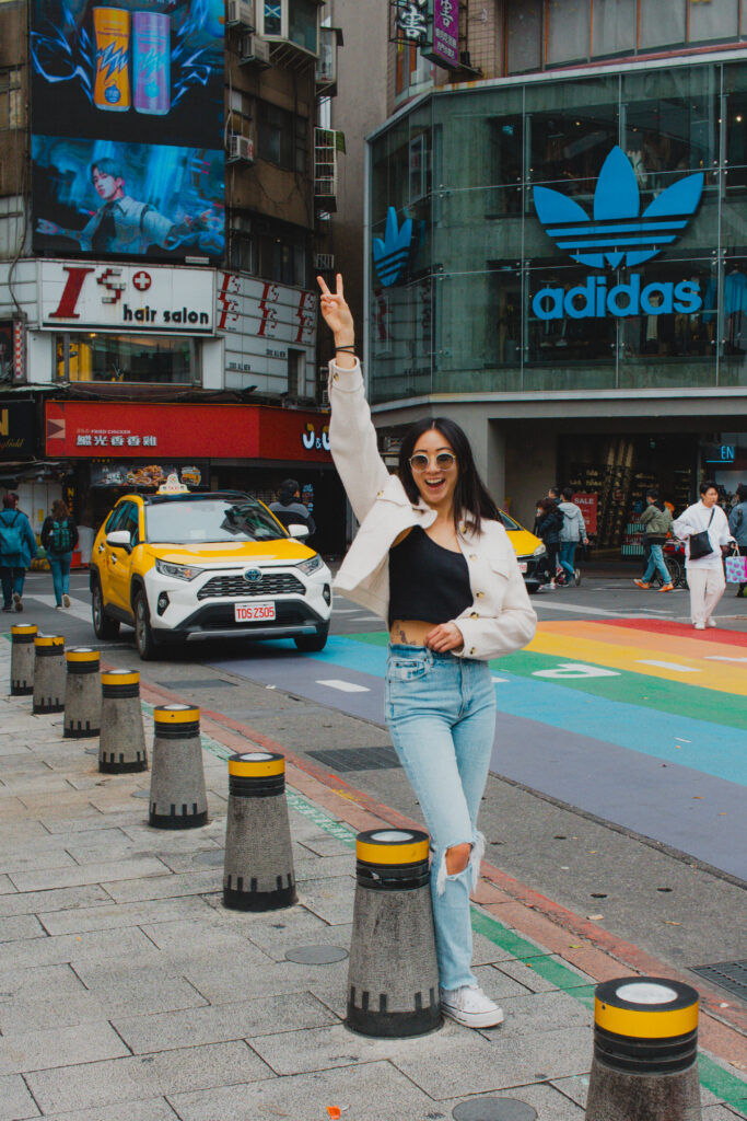 Smiling woman in a beige jacket raises her hand in a busy city street with a rainbow-colored crosswalk and an Adidas store in the background.