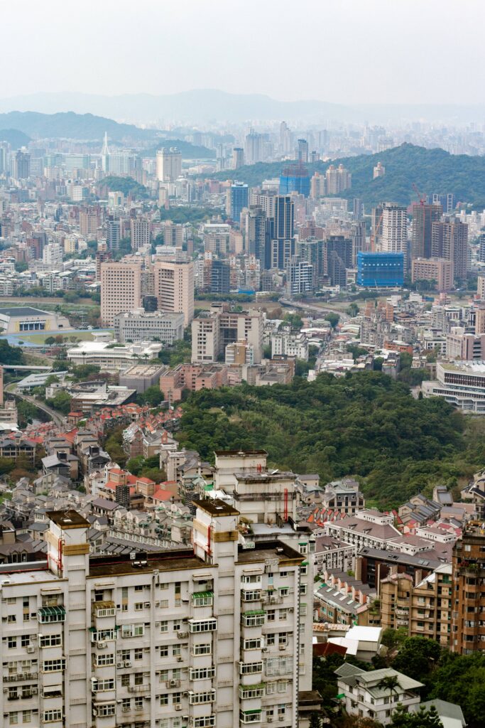 Wide cityscape with dense high-rise buildings and green hills in the background, under a pale sky.