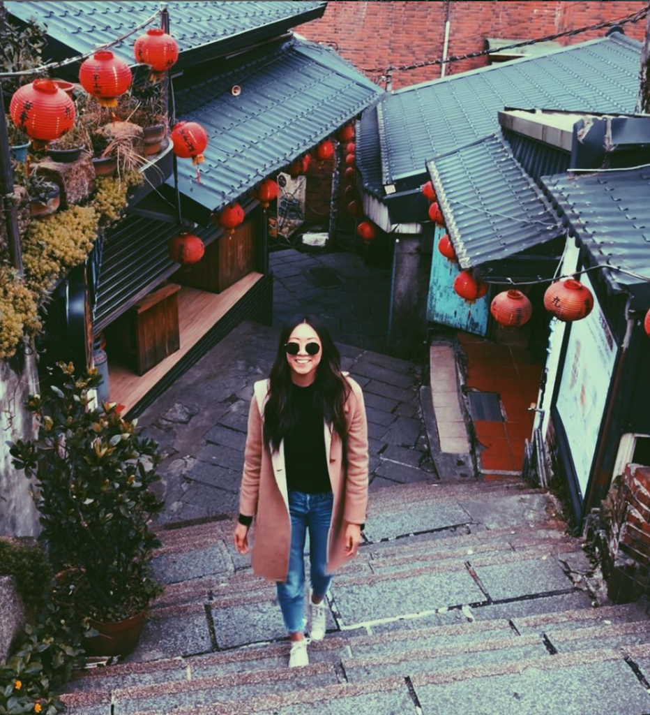 Woman walking up stone stairs in a narrow alley decorated with red lanterns, wearing a pink coat, black top, jeans and white sneakers.