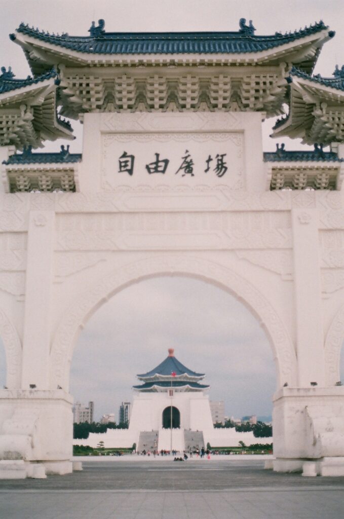 White ornate archway with blue-tiled Chinese roofs and a carved plaque with Chinese characters, opening to a distant pagoda-style building and courtyard.