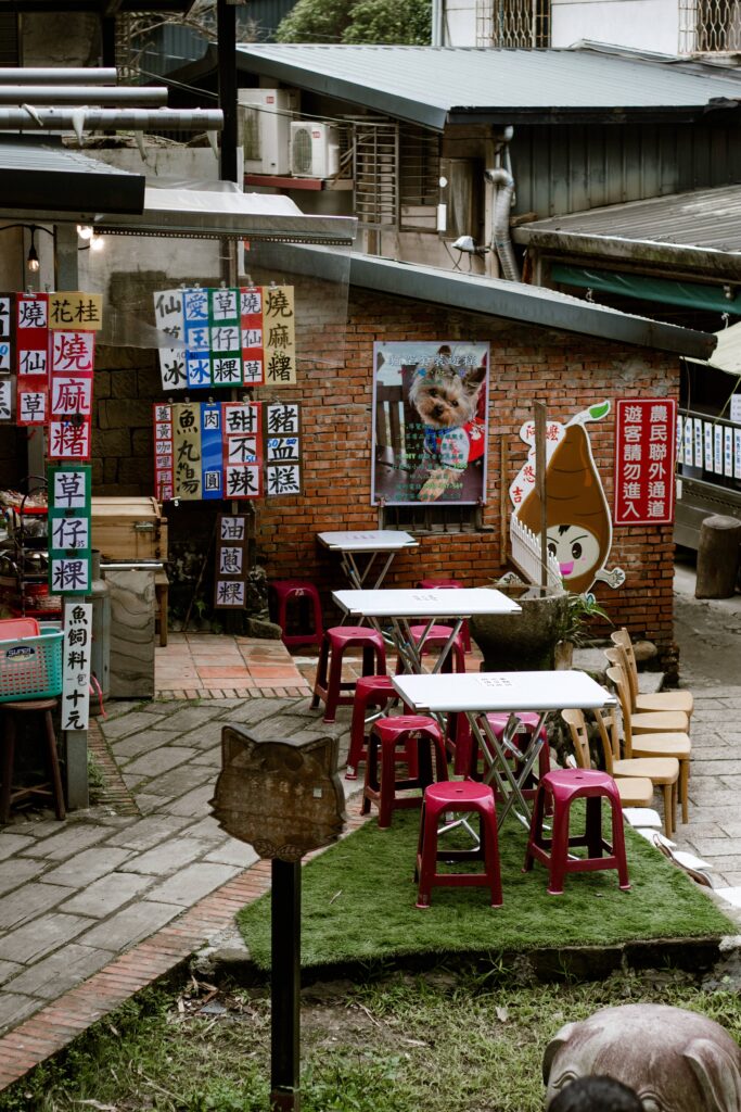 Outdoor alley dining area with red plastic stools and white tables against a brick wall, colorful signs with Chinese characters surrounding a poster of a dog/canine image.