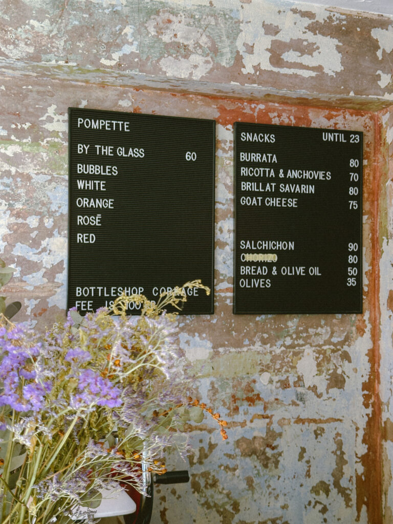 Two black menu boards mounted on a weathered, peeling wall; foreground shows purple dried flowers and greenery. (Wine by the glass, rosé, white, red listed)