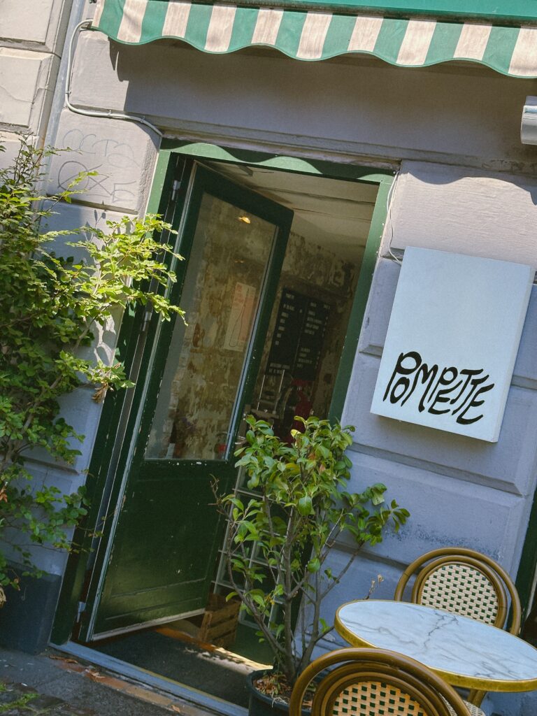 Open doorway of a small cafe with green door frame, striped green-and-white awning, and outdoor seating with marble-topped tables and wicker chairs surrounded by plants