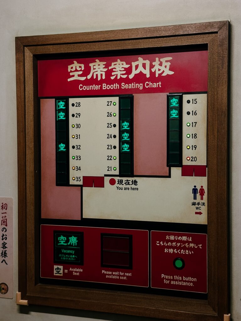 Framed red sign showing a counter booth seating chart with 'You are here' marker and green vacancy indicators, in Japanese and English text.