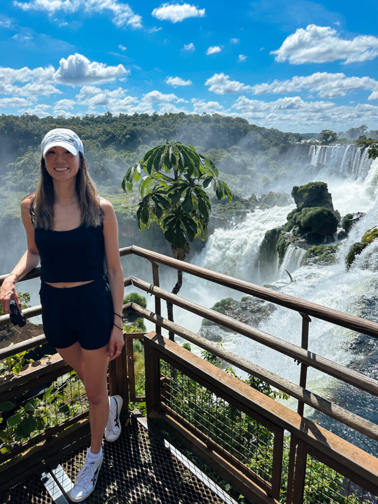 Woman in a white cap and navy outfit posing on a wooden observation deck with a massive waterfall and mist in the background.