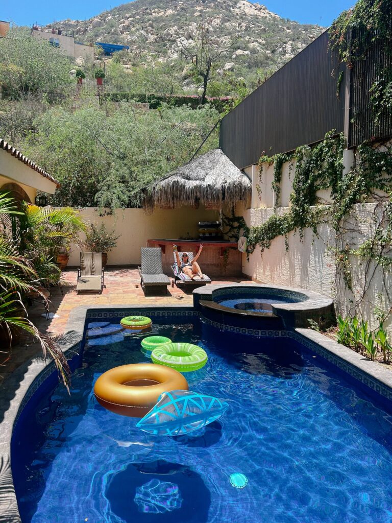 Backyard pool scene with blue water, several inflatable rings, and a person relaxing on a lounge chair under a thatched cabana.