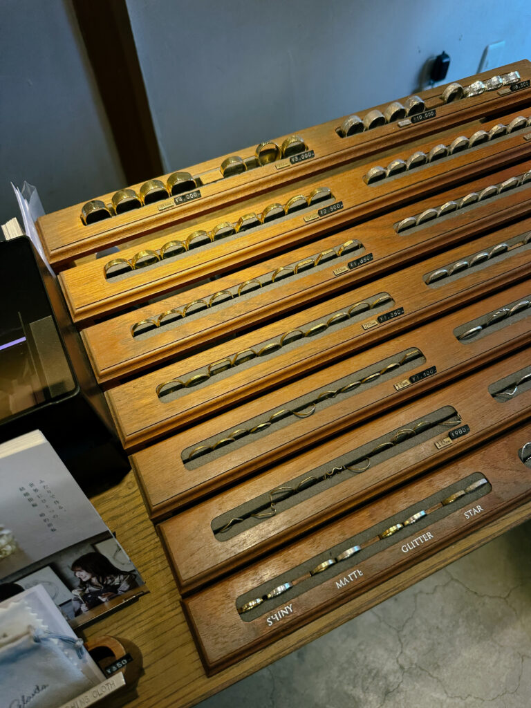 Wooden display trays holding rows of rings, organized by finish labels like Glitter, Shiny, Matte in a jewelry shop setting