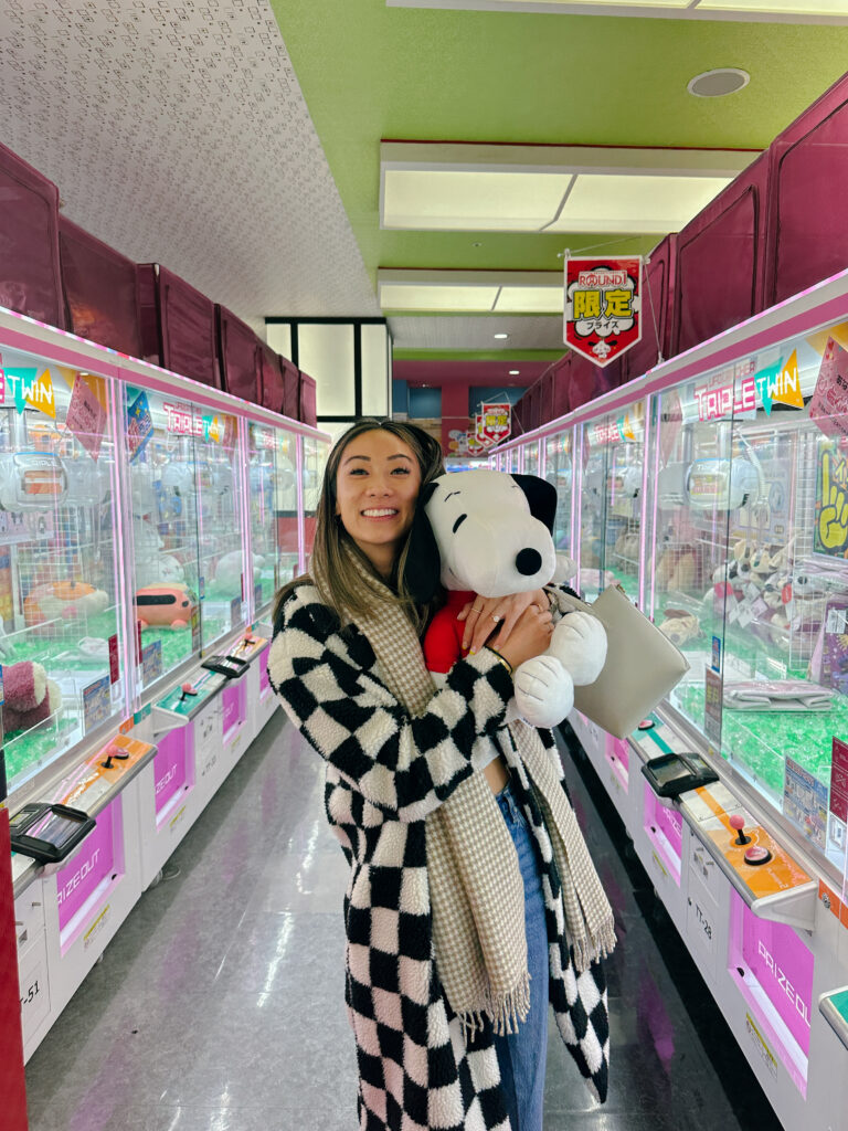 Smiling woman in a black-and-white checkered coat hugs a Snoopy plush in a pink arcade crane game hall.