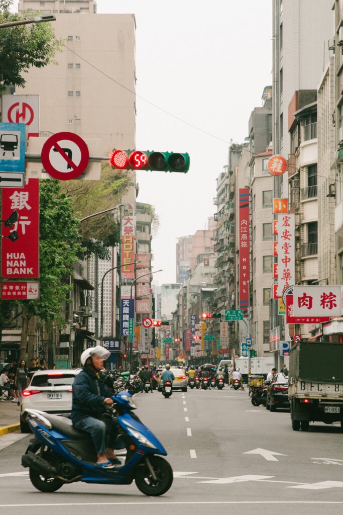 Busy urban street in an Asian city with scooters and cars, tall buildings, and red traffic lights overhead.