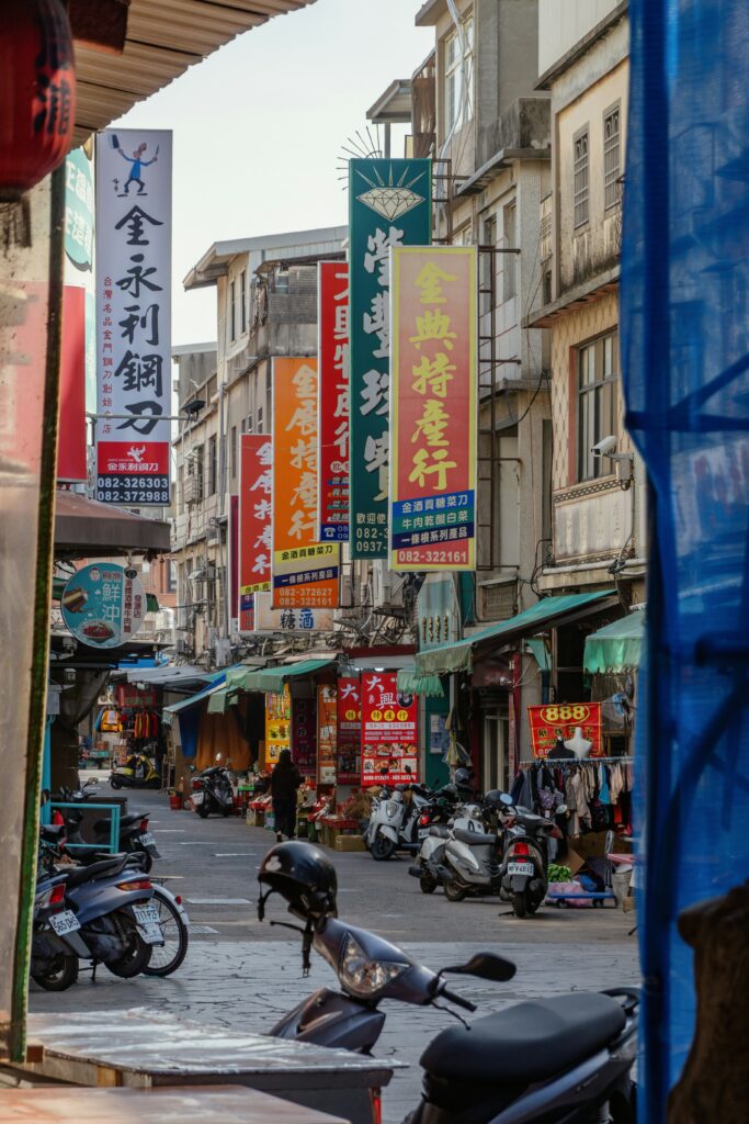 Busy street market lined with colorful vertical signs in Chinese characters and scooters parked along the curb.