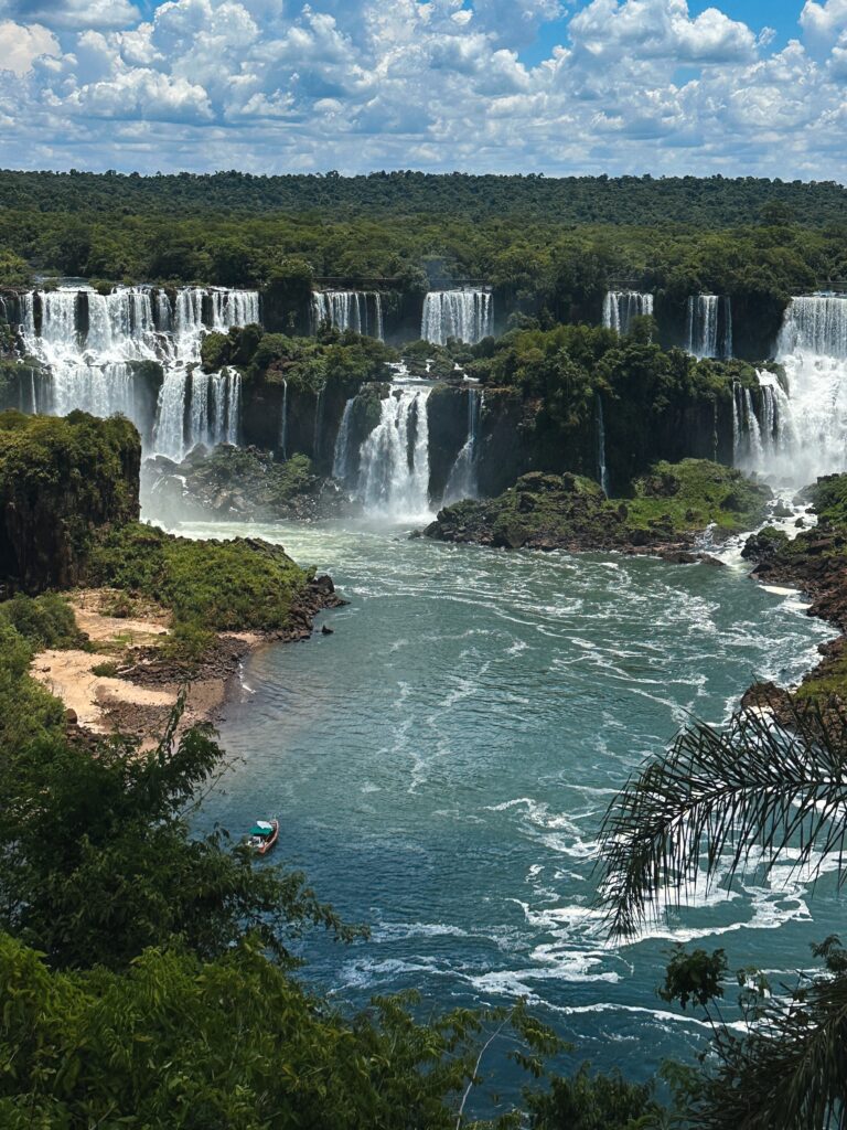 Wide view of multiple waterfalls spilling into a wide river, surrounded by dense green forest and rocky cliffs with mist in the air.