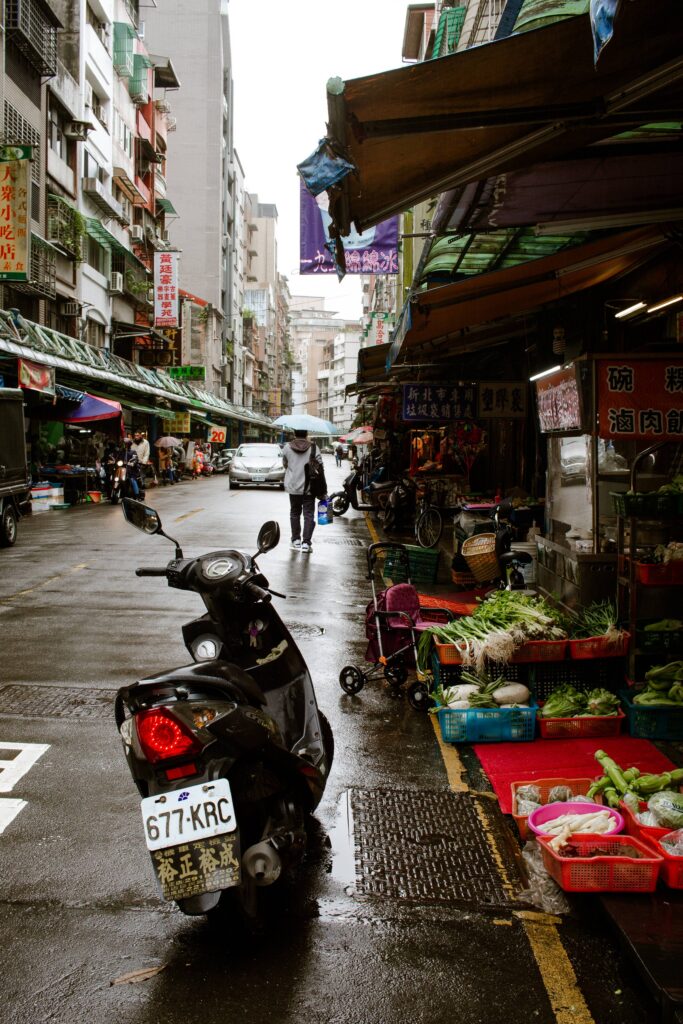 Narrow, wet city street market in Asia with vegetable stalls on the right and scooters parked along the curb; tall buildings with signs on the left.