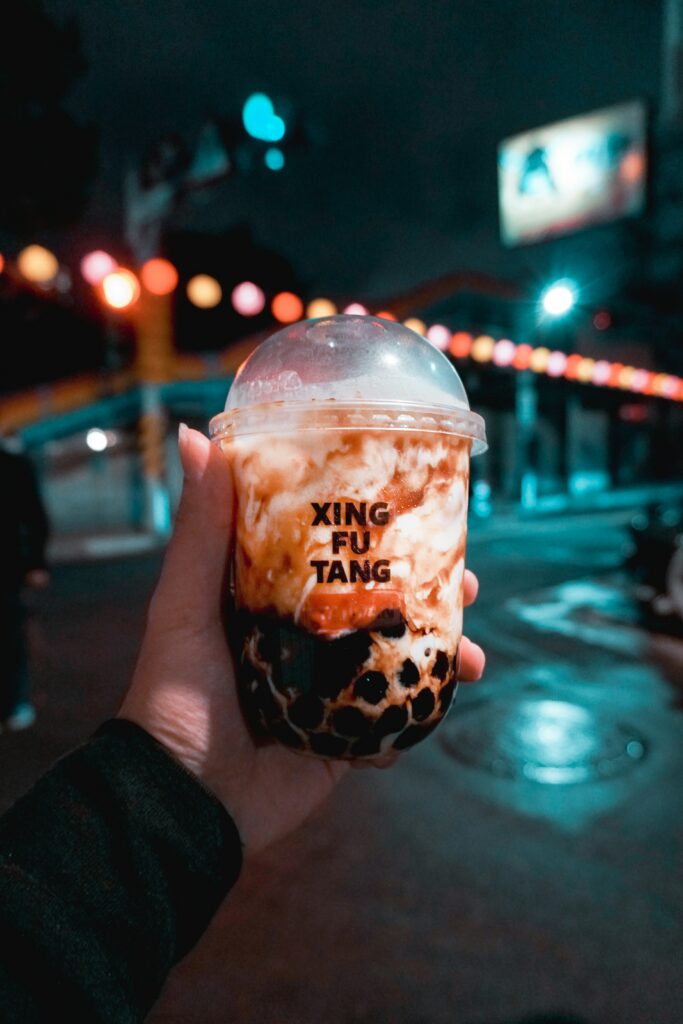 Hand holding a cup of bubble tea with tapioca pearls in the foreground, colorful night lights in the background and a brand name on the cup read as XING FU TANG