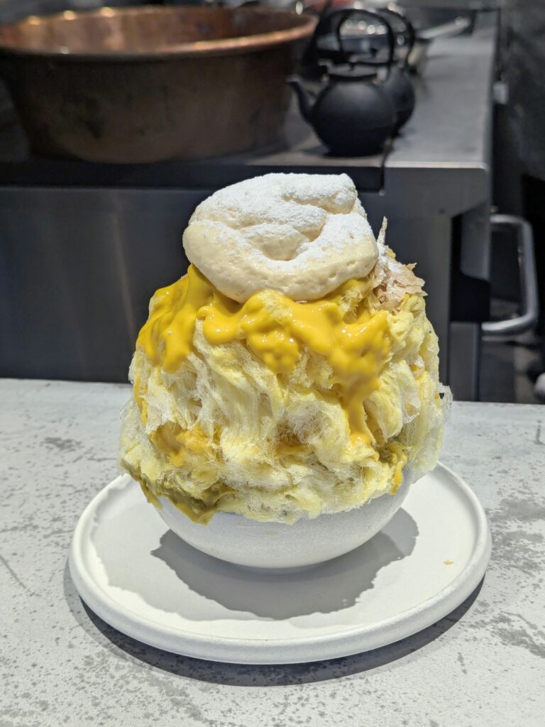 Taiwanese Shaved-ice dessert with orange syrup in a dark bowl on a wooden tray, with side bowls of colorful mochi and toppings nearby.