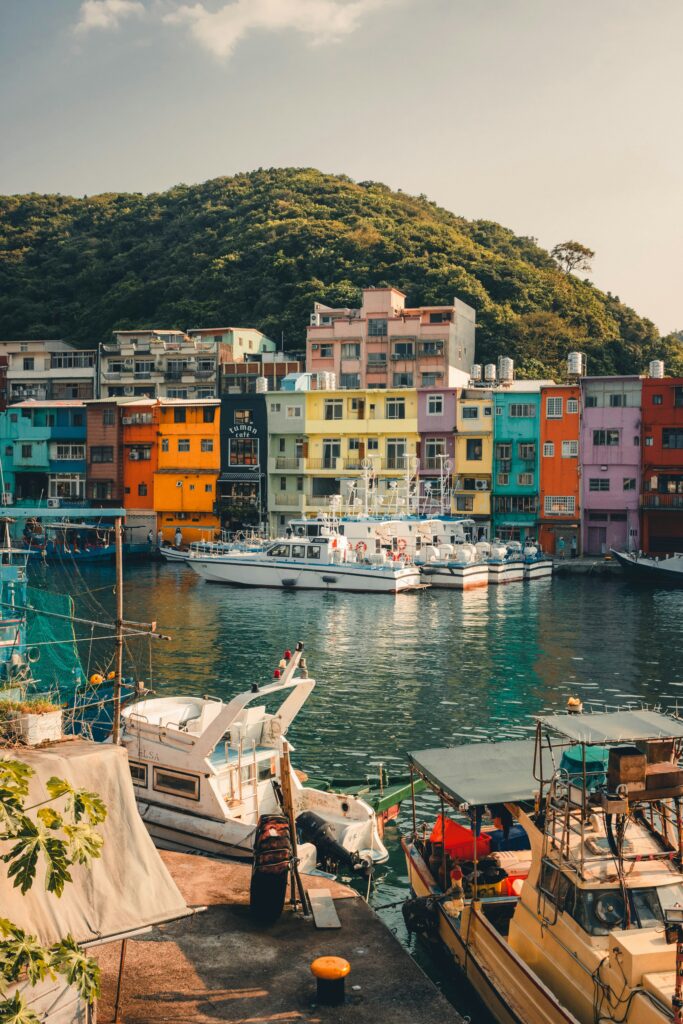 Colorful row of houses lining a marina with docked boats and a green hillside in the background.