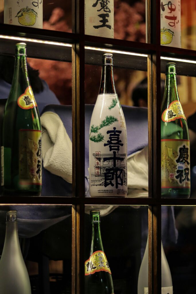 Row of sake bottles on a wooden shelf with Japanese labels, center bottle white with green pine design.