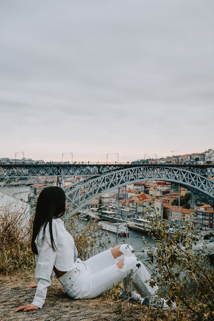 girl sitting in front of porto bridge deciding between visiting lisbon vs porto